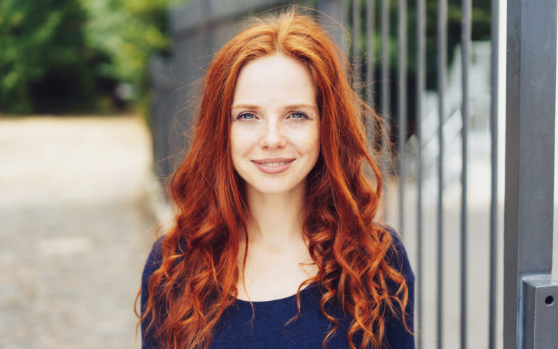 Pretty young woman with gorgeous long curly red hair standing outdoors alongside a metal railing looking at the camera with a quiet thoughtful friendly smile
